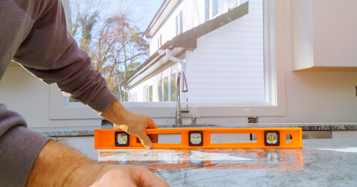 A man holds an orange level against a gray granite countertop across from a kitchen sink and window.