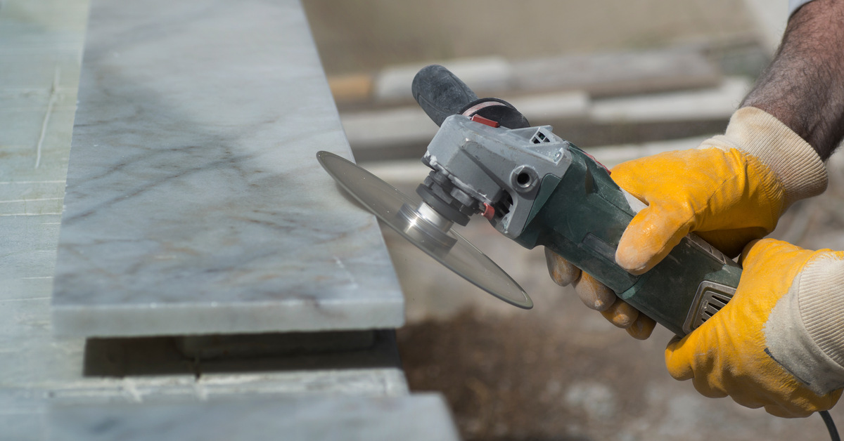 Someone wearing yellow work gloves holds a grinder at an angle against a small slab of marble resting on a work table.