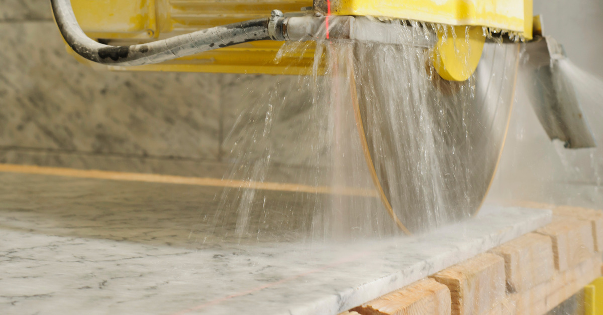 A large flat piece of marble rests on two-by-fours. A large saw with water spraying over the stone cuts the material.