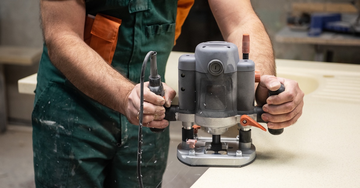 Someone wearing an orange t-shirt and green coveralls holds a grinder against the edge of a countertop.