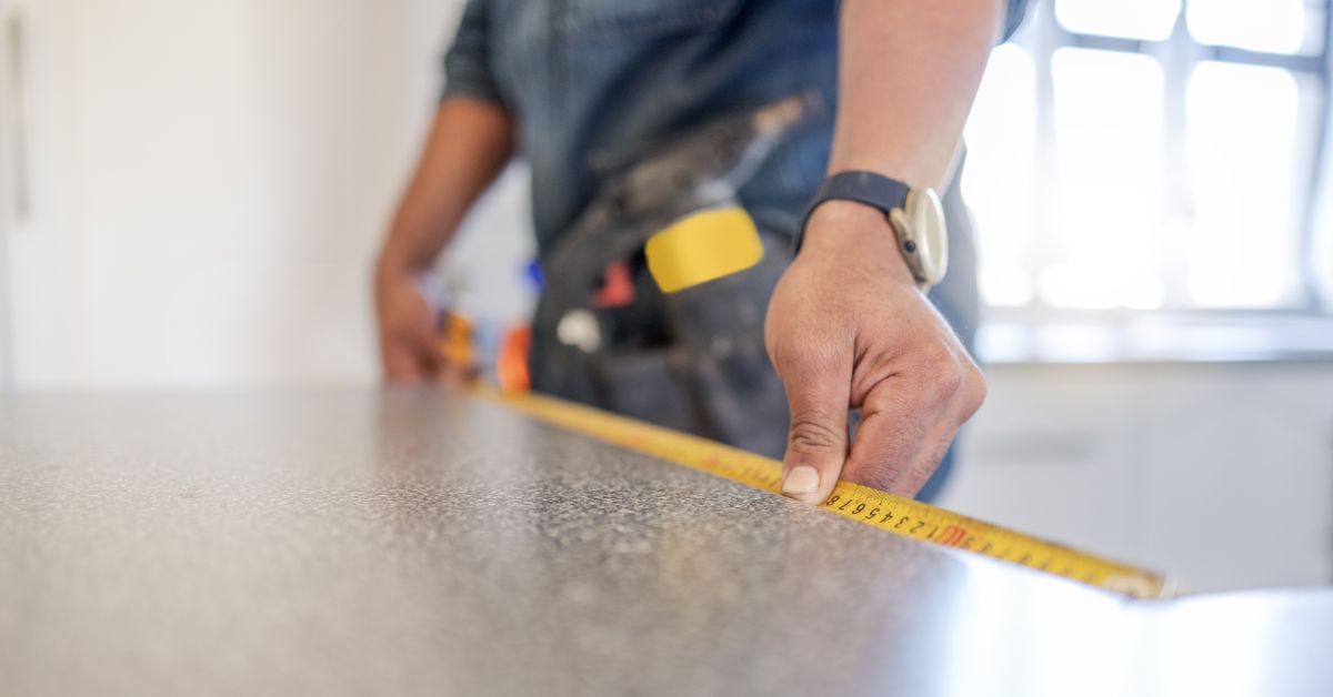 A close-up of someone holding a tape measure against the edge of a counter. A window behind the person brings in light.