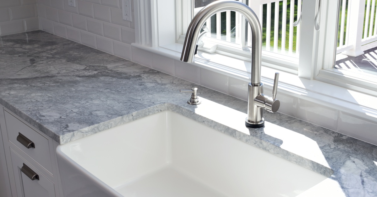 A porcelain farmhouse kitchen sink in front of a sunny window. The countertops are made from grey marble.