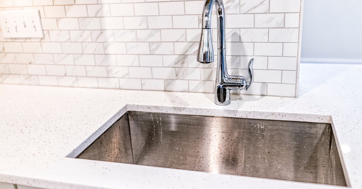 A kitchen with a stainless steel undermount sink and white granite countertops. The backsplash is also made of white marble tiles.