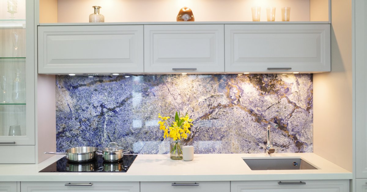 White kitchen cabinets with white counters are brightly lit. The backsplash features a large slab of marble.