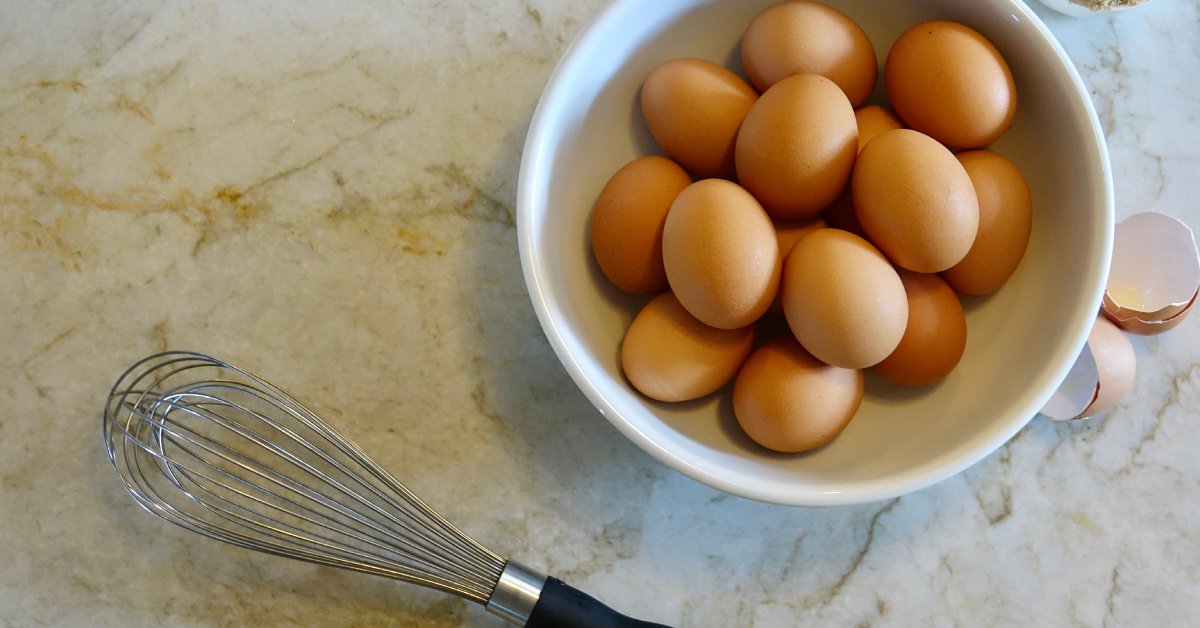 A large ceramic bowl of whole eggs, a whisk, and a small ceramic bowl of brown sugar on top of a quartzite counter.